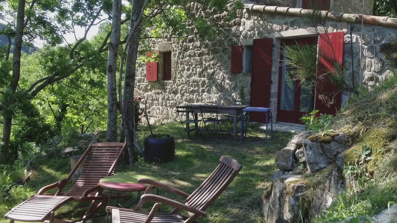 La terrasse de la maison, ses transats à l'ombre des arbres et la porte-fenêtre aux volets rouges